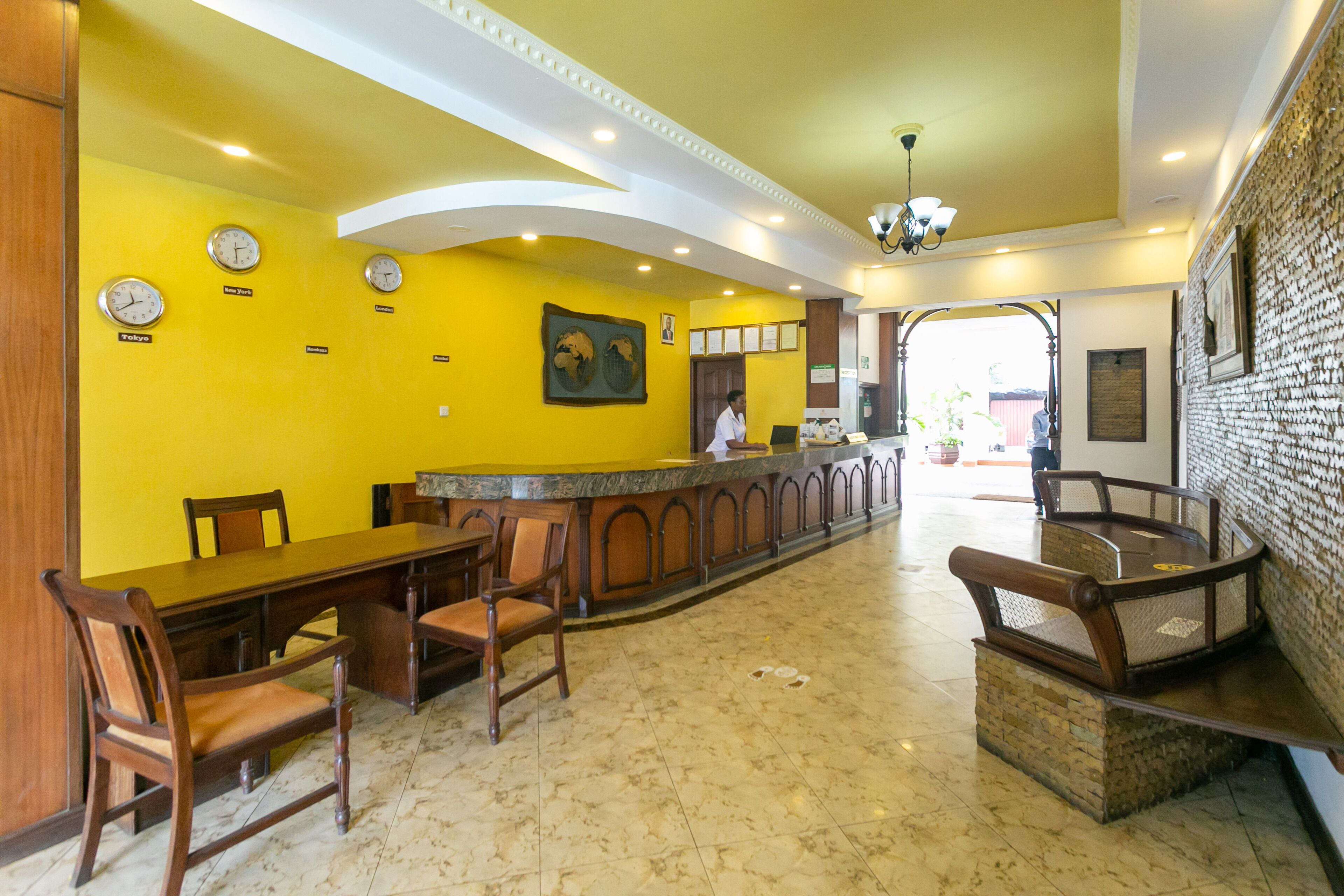 A restaurant with yellow walls, clocks indicating different time zones, a long wooden table, and a counter with a person behind it.