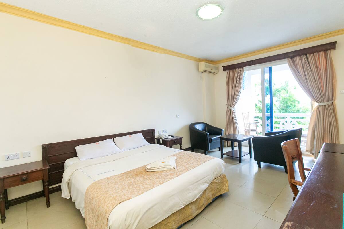 A hotel room with a large bed, a desk, a chair, and a balcony with curtains.