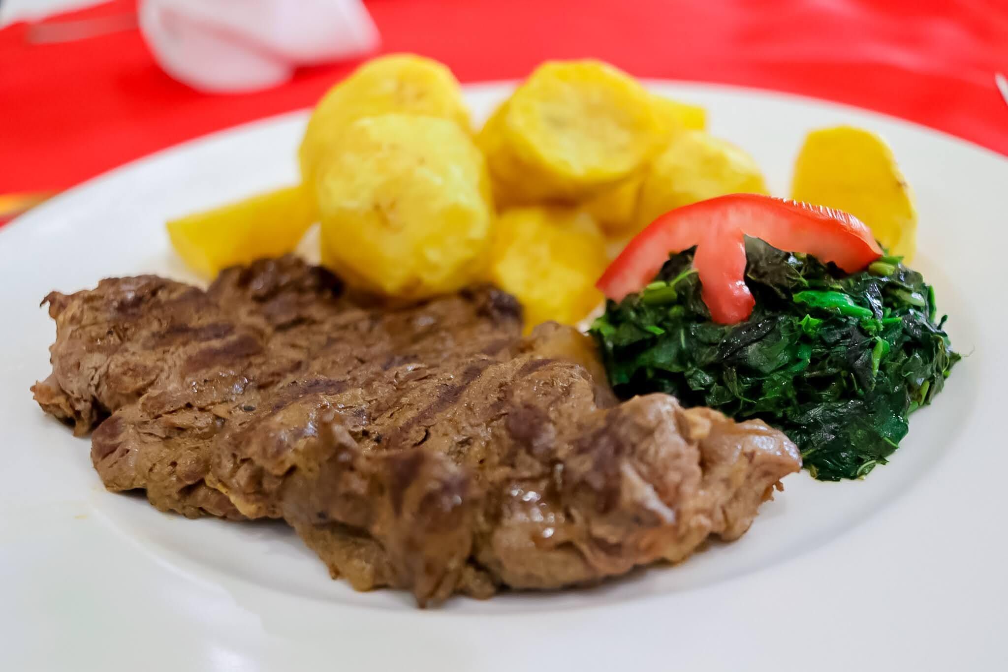 Un plato con una porción de carne, patatas amarillas y verduras de hoja verde.