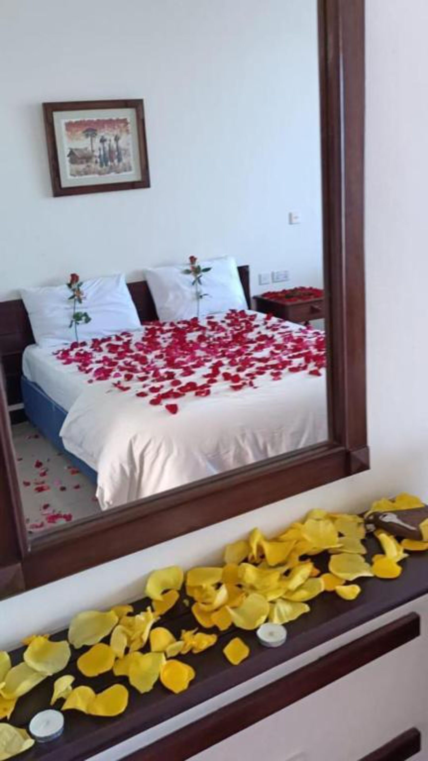 A bed with rose petals and a candle on a dresser.