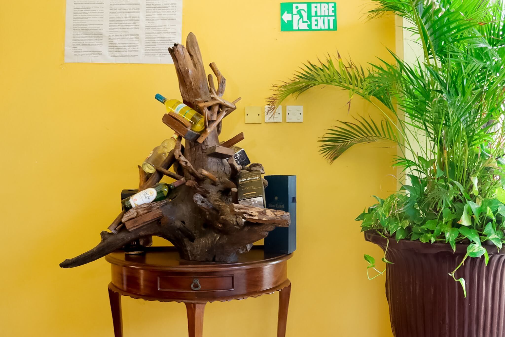 Eine Holzschnitzerei mit Flaschen auf einem Tisch vor einer gelben Wand, ein Feuerschutzschild und ein Blumentopf.