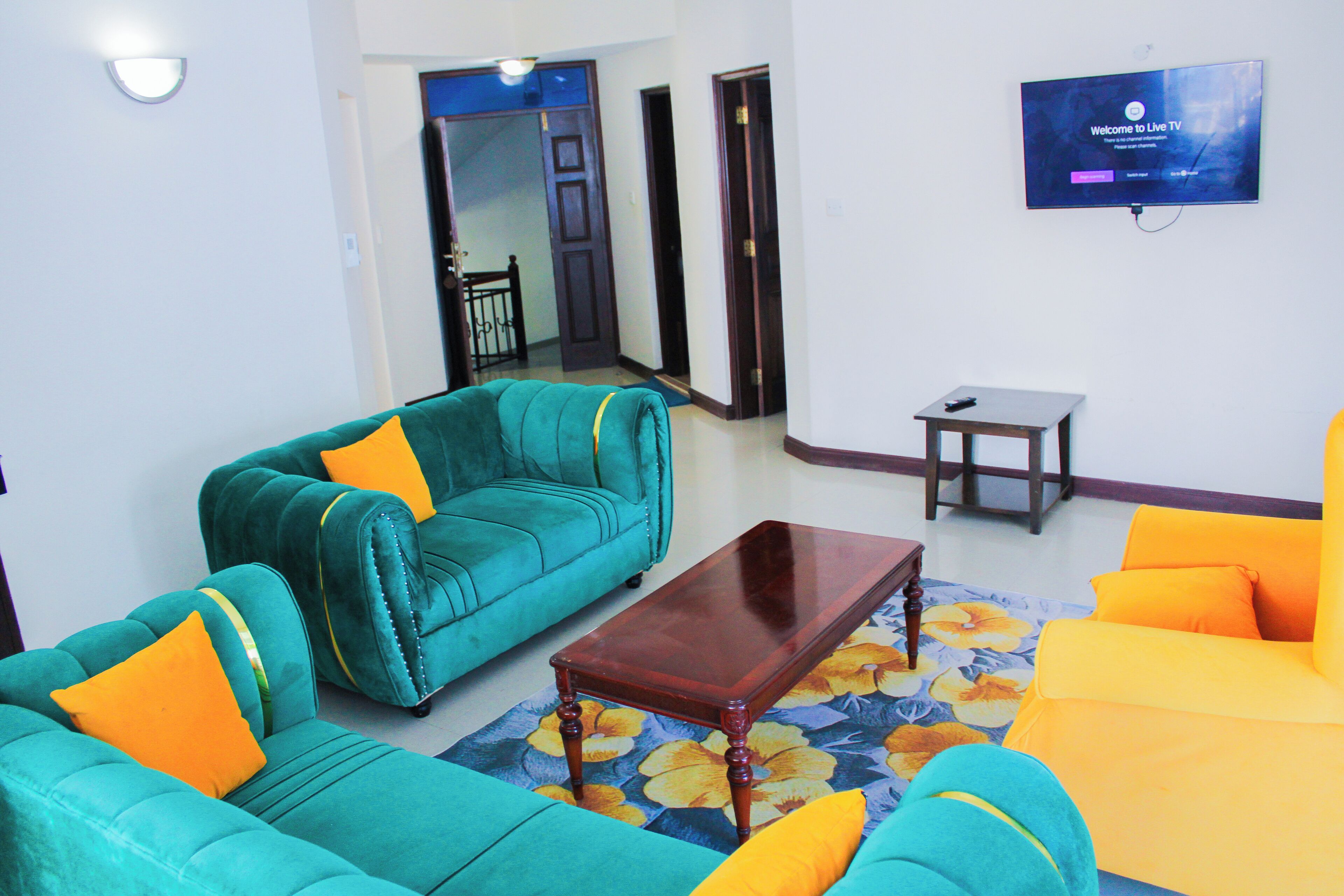 A living room with teal and orange sofas, a wooden coffee table, and a TV on the wall.