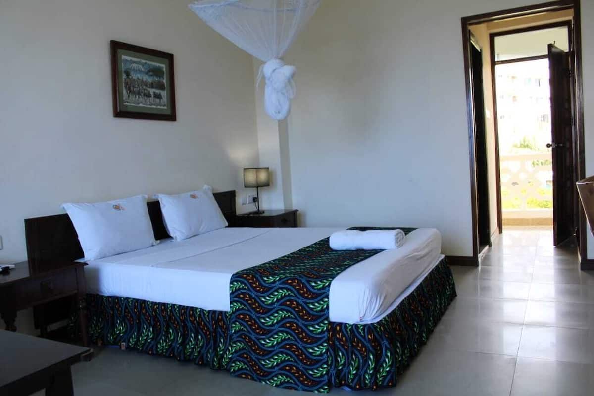 A hotel room with a bed, a bedside table, a lamp, a framed picture on the wall, and a door leading to an outdoor area.