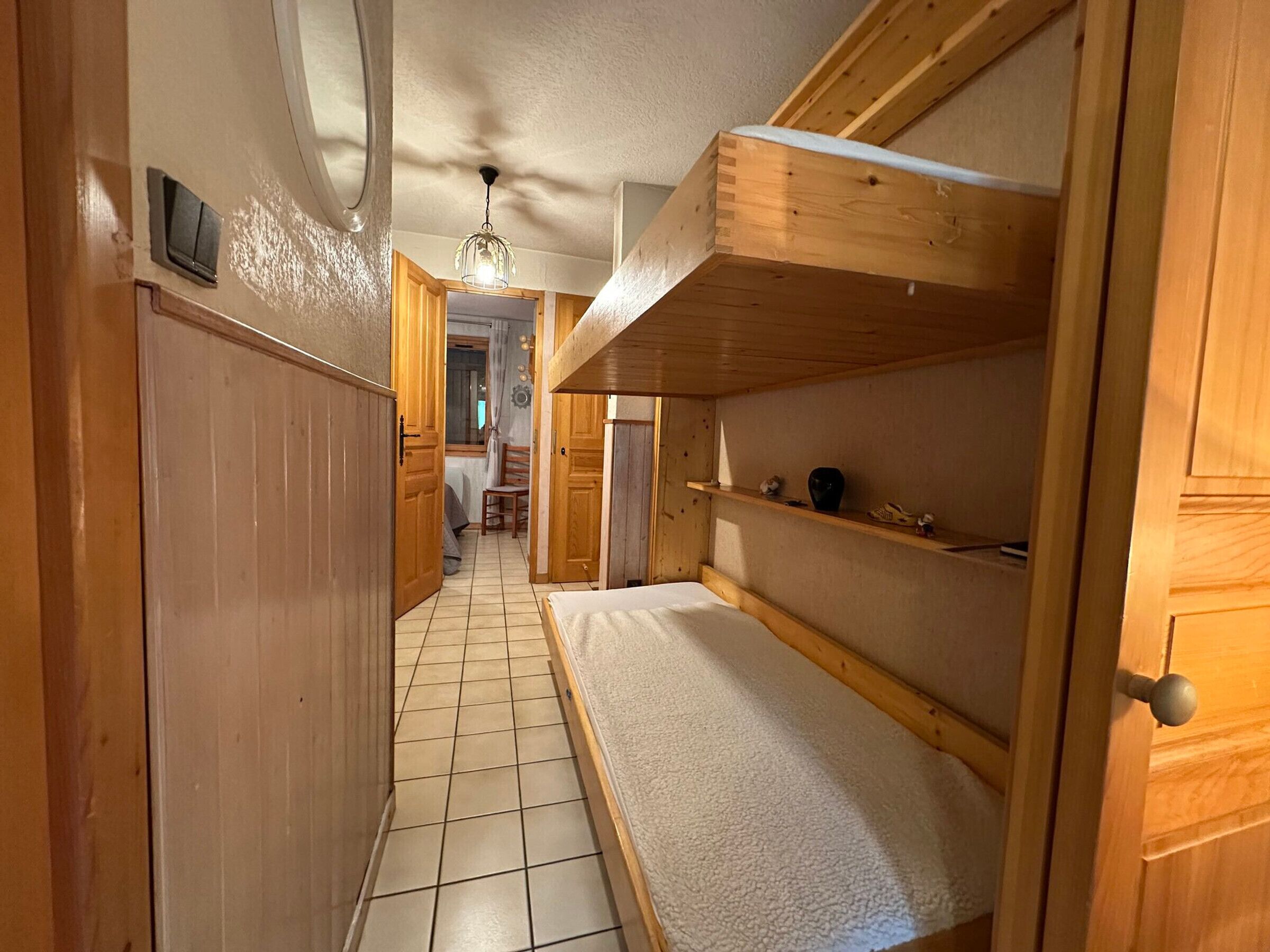 A narrow hallway with wooden walls and a tiled floor. There is a bed with a headboard and a shelf above it. A door is visible on the left, and a window is seen in the background.