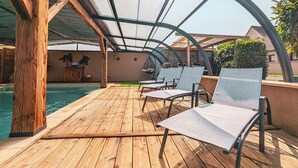 Een overdekt buitengebied met een zwembad, een houten terras en lounge stoelen.
