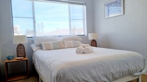 A neatly made bed with pillows and rolled towels, a bedside lamp, and a window with a view of buildings. - Welcome to Champagne Court 07 (Tuncurry)