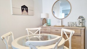 A dining area with a round glass table, white chairs, a wooden cabinet with decorative items, a framed picture, and a mirror on the wall. - Welcome to Champagne Court 07 (Tuncurry)