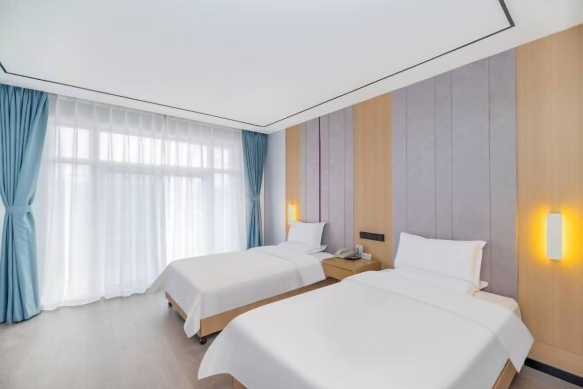 A hotel room with two beds, a nightstand, a bedside lamp, and a large window with curtains.
