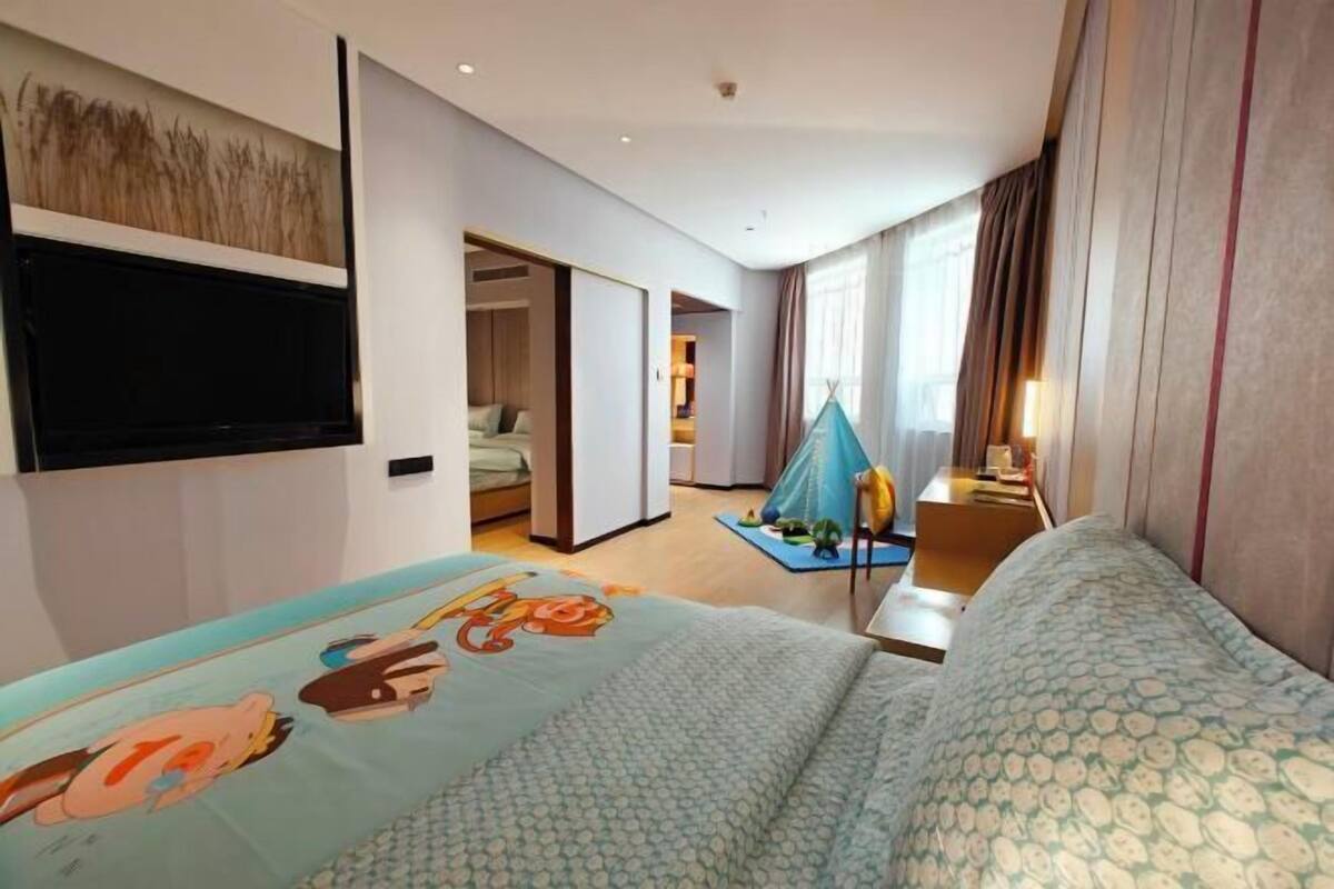 A hotel room with a bed, a desk, a television mounted on the wall, and a large window with curtains.