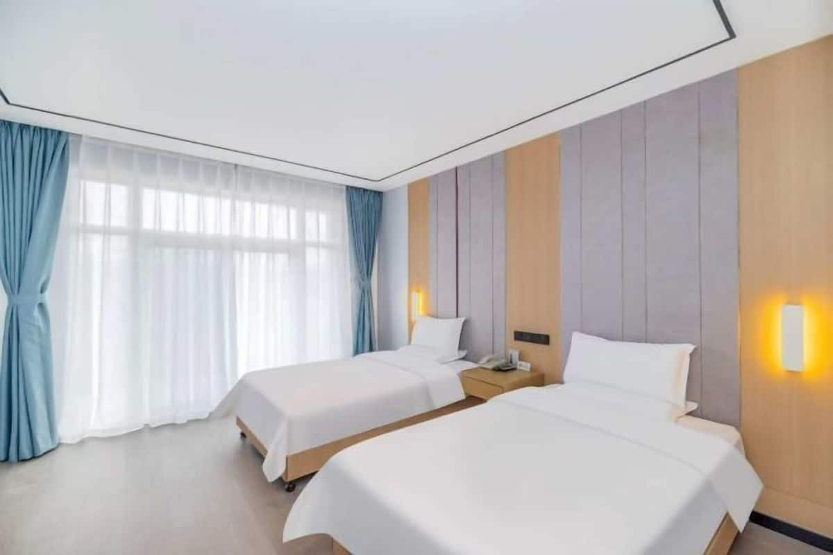 A hotel room with two beds, a large window with curtains, and a wall-mounted light fixture.