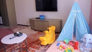 A room with a television mounted on the wall, a small round table, a yellow toy, a blue play tent, and various children's toys. - RGHCM Garden Resorts (Beijing)
