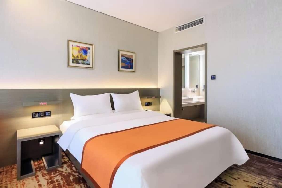 A hotel room with a bed, nightstands, a digital clock, artwork on the wall, and a mirror.