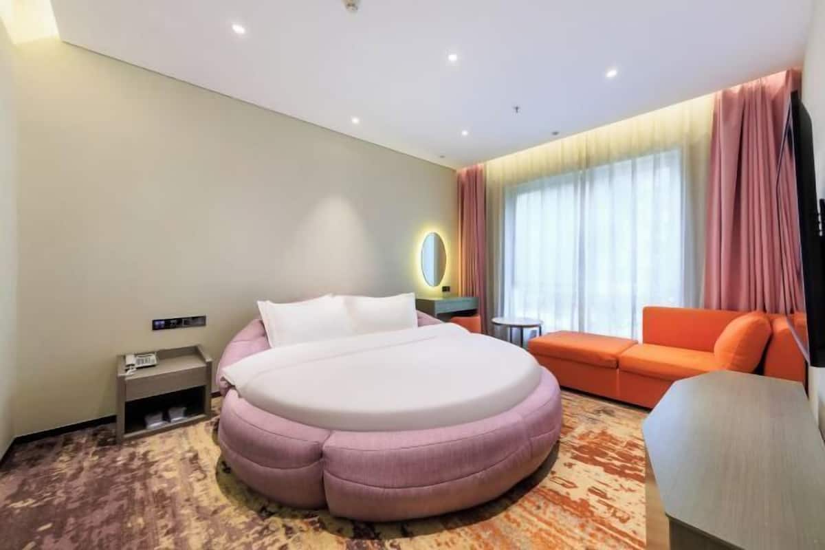 A modern hotel room with a round bed, an orange sofa, a small side table, and a large window with curtains.