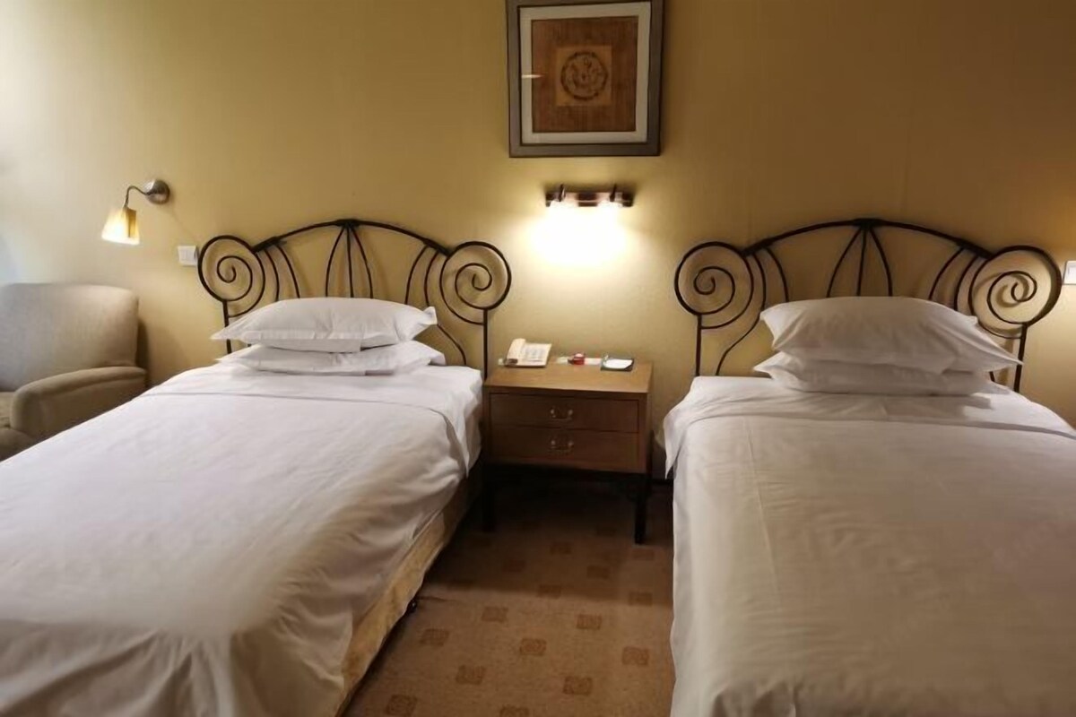 A hotel room with two single beds, a nightstand between them, a chair, and a wall-mounted light fixture.