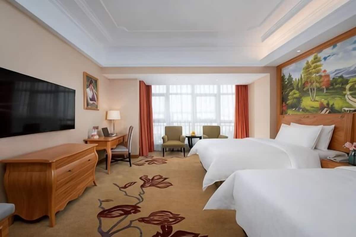 A hotel room with a large bed, a smaller bed, a desk with a chair, a TV, and a painting on the wall.