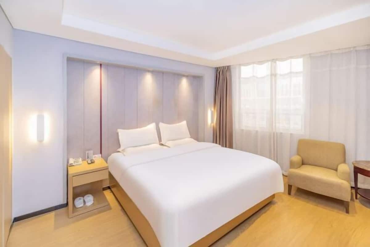 A hotel room with a large bed, a bedside table with a lamp, a chair, and a window with curtains.