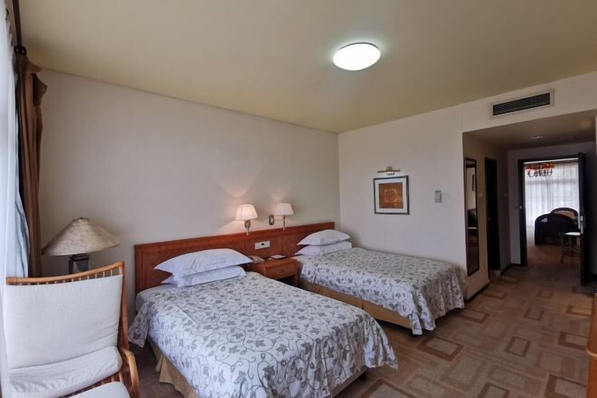 A hotel room with two beds, a wooden headboard, a chair, a lamp, and a framed picture on the wall.