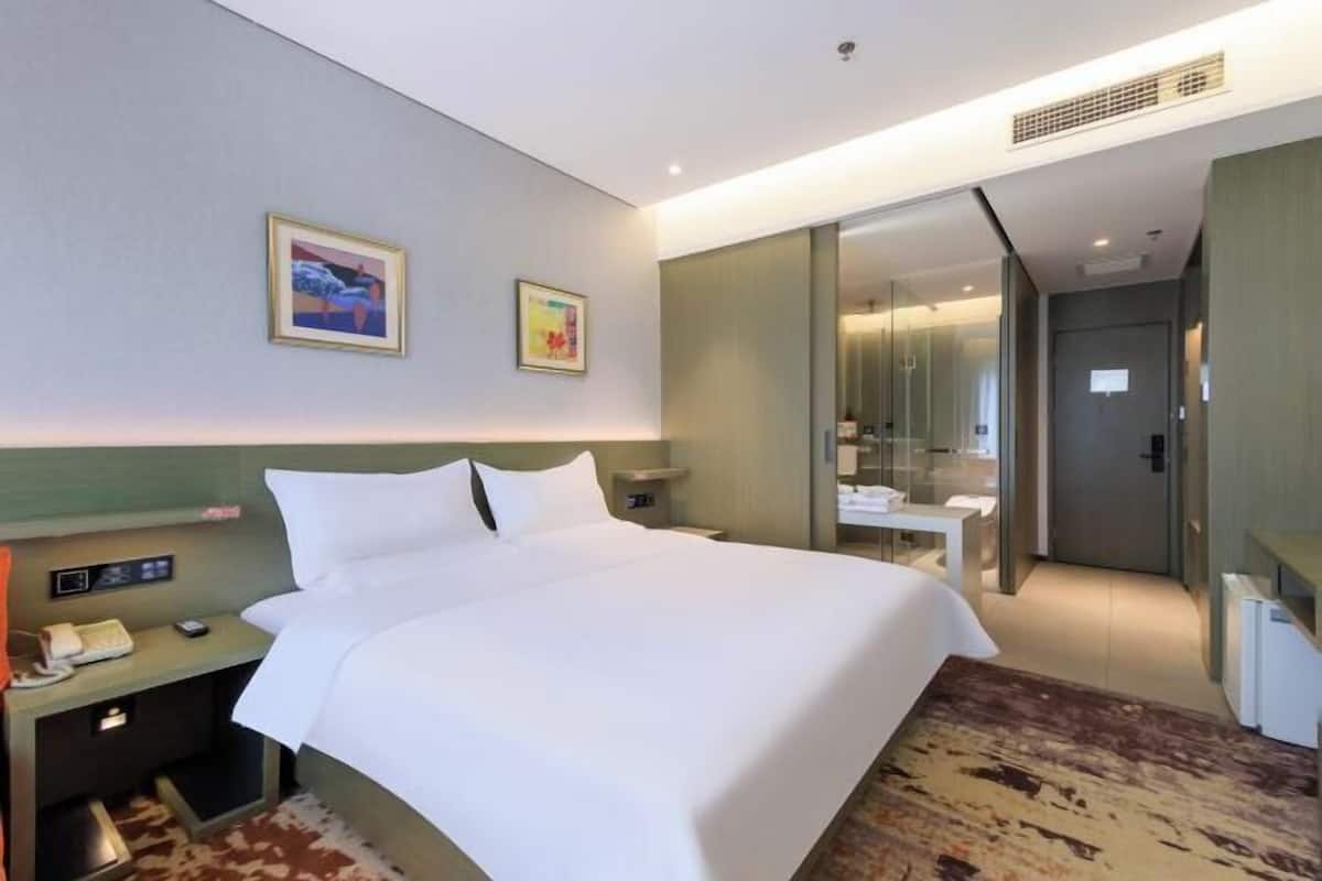 A hotel room with a large bed, bedside tables, a desk, a mirror, and a bathroom visible through a glass partition.