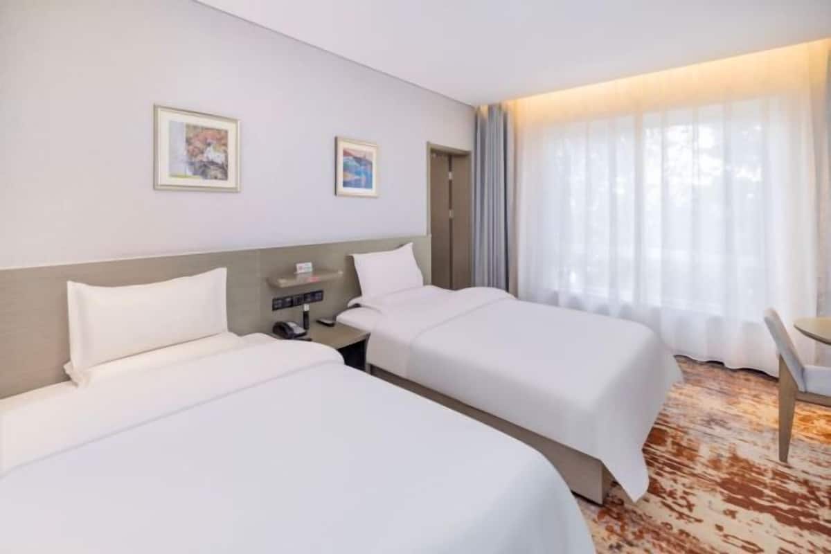 A hotel room with two beds, a bedside table, a chair, and a large window with curtains.