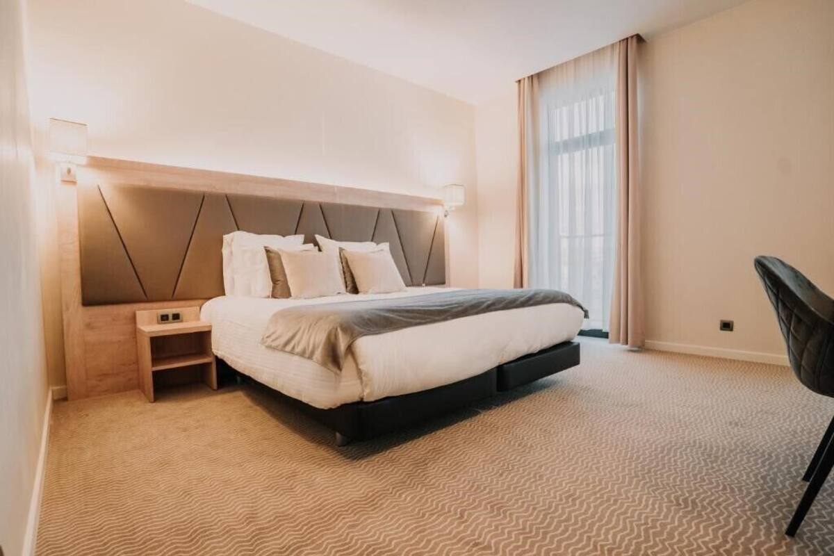 A modern hotel room with a bed, bedside tables, and a large window with curtains.