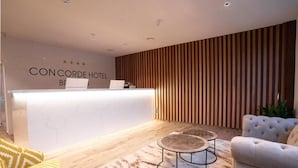 Zona de recepción de un hotel con un mostrador blanco, un panel de listones de madera y un área de descanso con dos mesas redondas y un sofá.