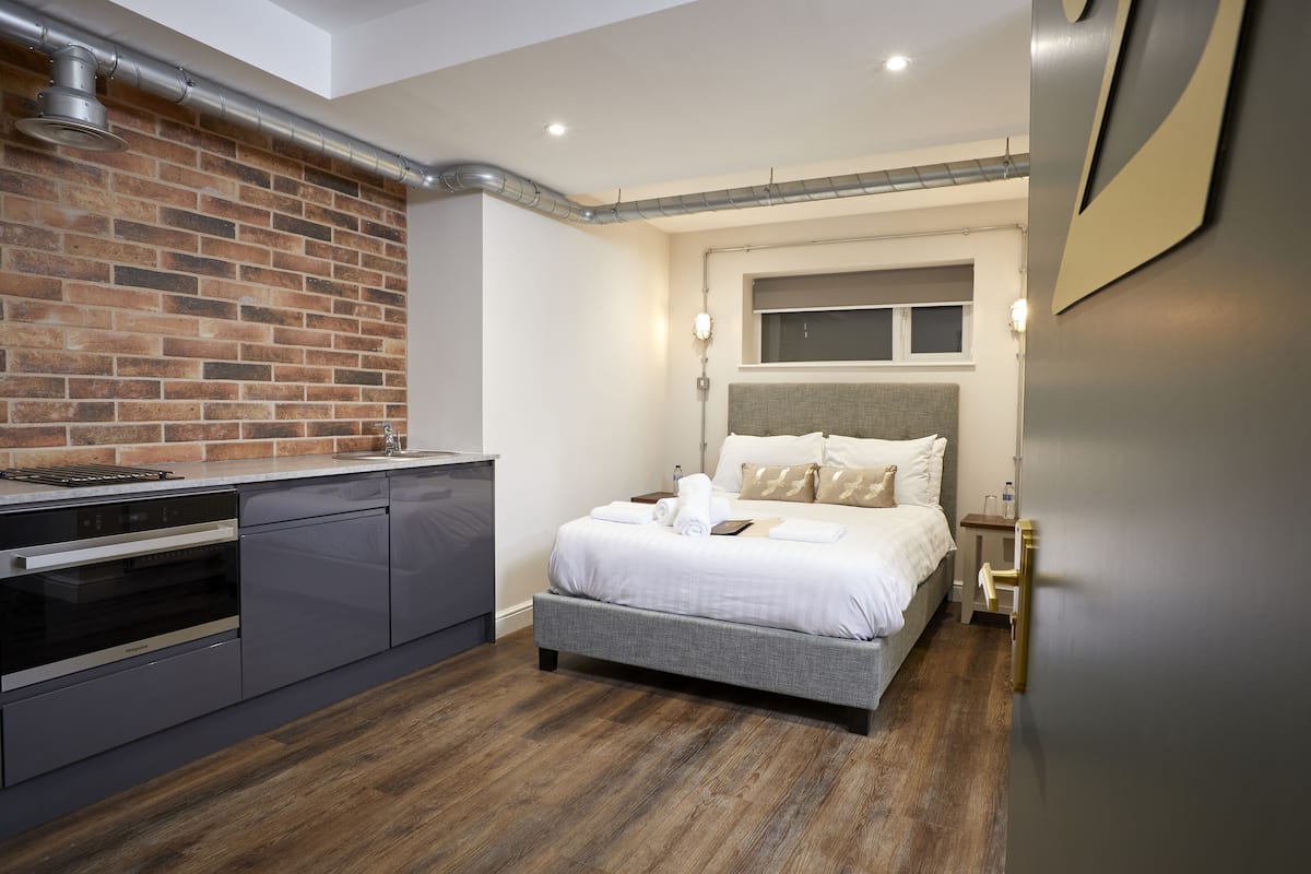 A modern bedroom with a brick accent wall, a built-in kitchen with a dishwasher, a grey bed with white bedding, and a bedside table with a lamp.