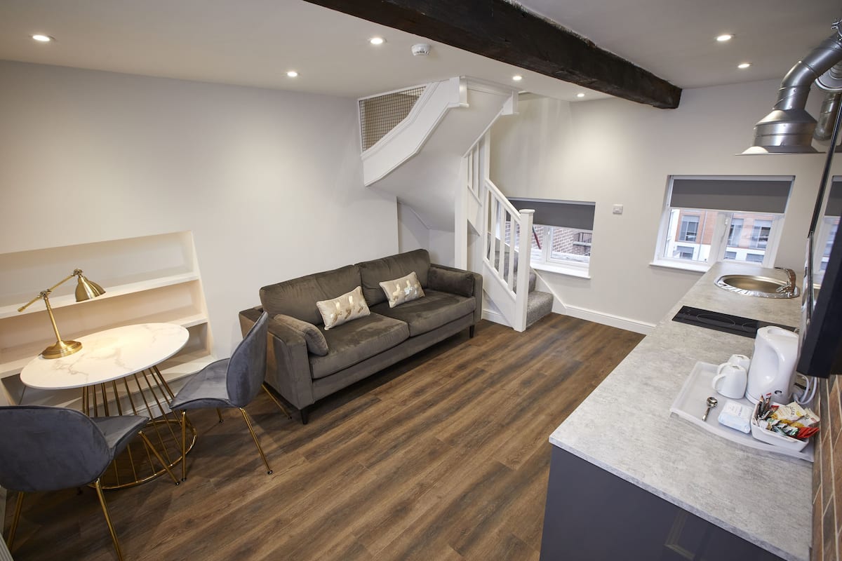 A modern living room with a grey sofa, a round coffee table, and a dining area with a white table and chairs. A staircase leads to an upper level.