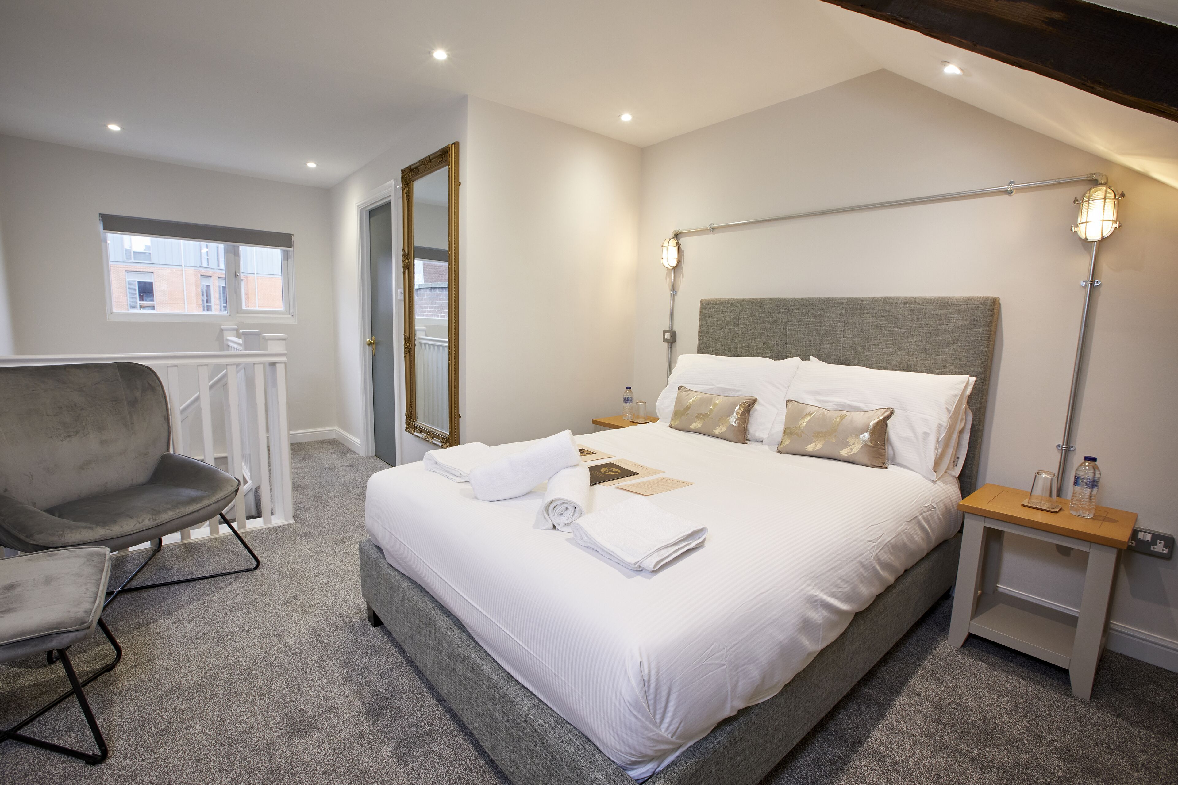 Een keurig opgemaakt bed met witte lakens, een grijs gestoffeerde fauteuil en een nachtkastje met een lamp.