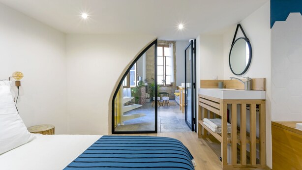 A bedroom with a bed, a wooden bedside table, a mirror, and a view into a living area with a sofa and a table. - L'Esprit Montpel "Face à l'Eglise St Roch" (Montpellier Spirit) (Montpellier)