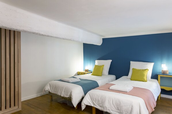 A room with two beds, each with a white bedspread and a colorful pillow. There are wooden floors and a white ceiling with exposed beams.