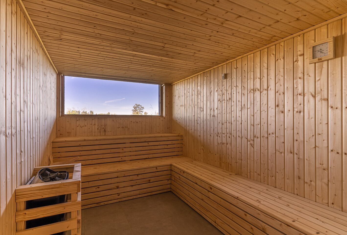Un sauna aux murs et aux bancs en bois, une fenêtre donnant sur les arbres et une horloge au mur.
