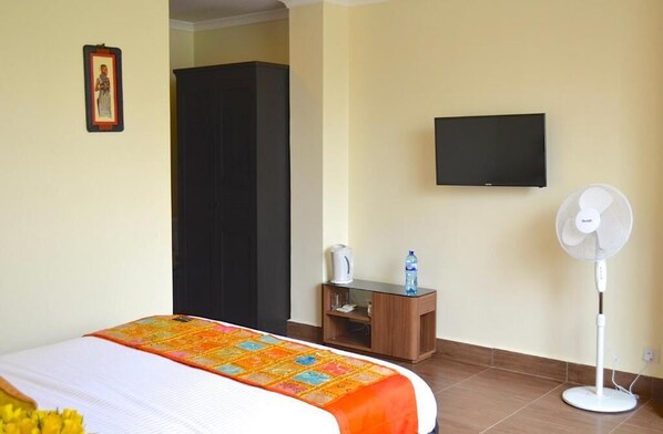 A hotel room with a bed, a television mounted on the wall, a standing fan, and a small table with a kettle and a bottle. - Gigiri Express Hotel (Nairobi)