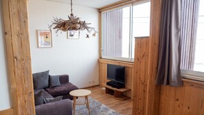 A living room with a wooden floor, a purple sofa, a small round table, a TV on a wooden stand, and a window with curtains.