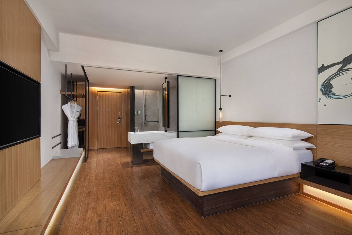 A modern hotel room with a large bed, a built-in TV, a bathroom with a mirror, and wooden flooring.