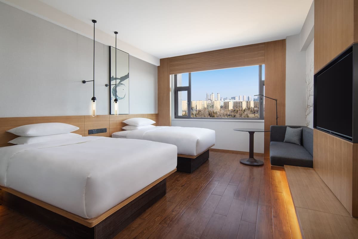 A modern hotel room with two beds, a sofa, a TV, and a large window with a city view.