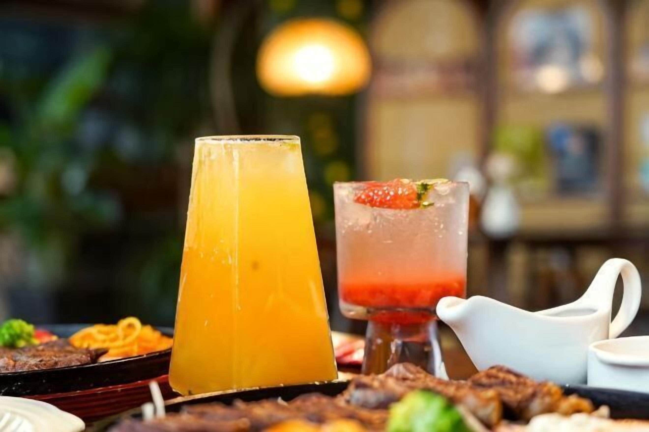 A table with a tall glass of orange juice, a glass with a red drink and a strawberry garnish, and a white pitcher.
