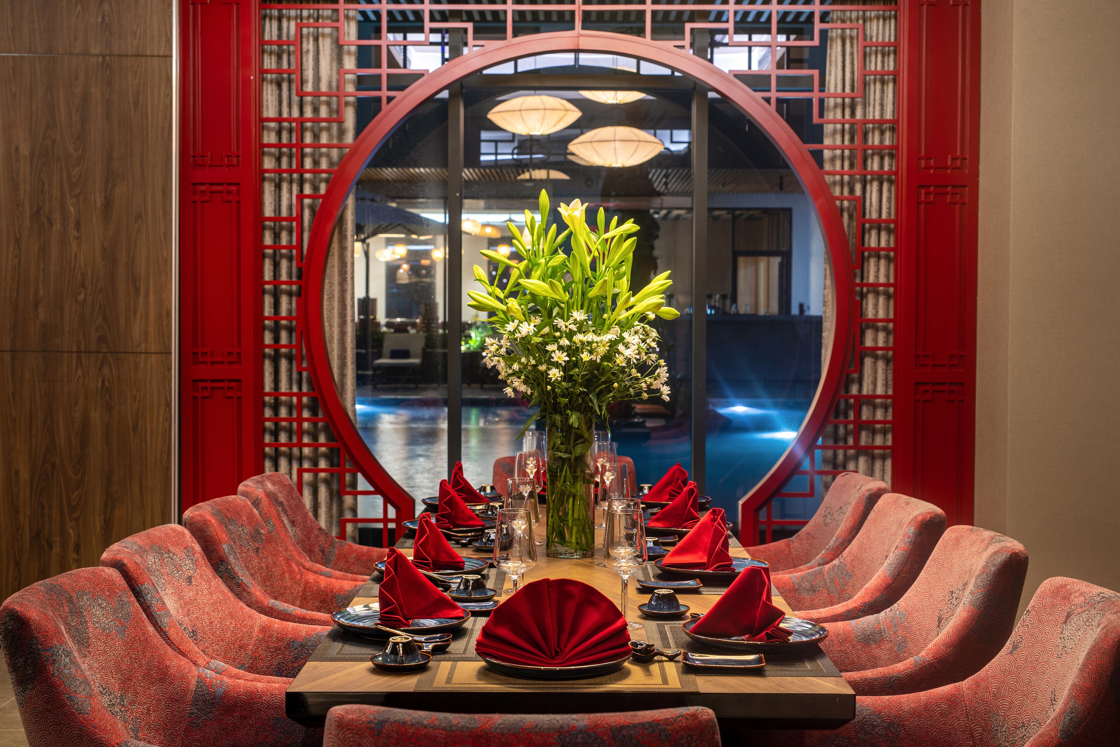 Mesa de comedor dispuesta con servilletas rojas, un jarrón de flores y vista a un cuerpo de agua a través de un ventanal circular.