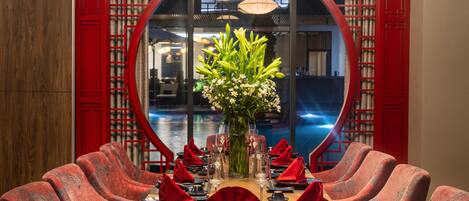 Mesa de comedor dispuesta con servilletas rojas, un jarrón de flores y vista a un cuerpo de agua a través de un ventanal circular.