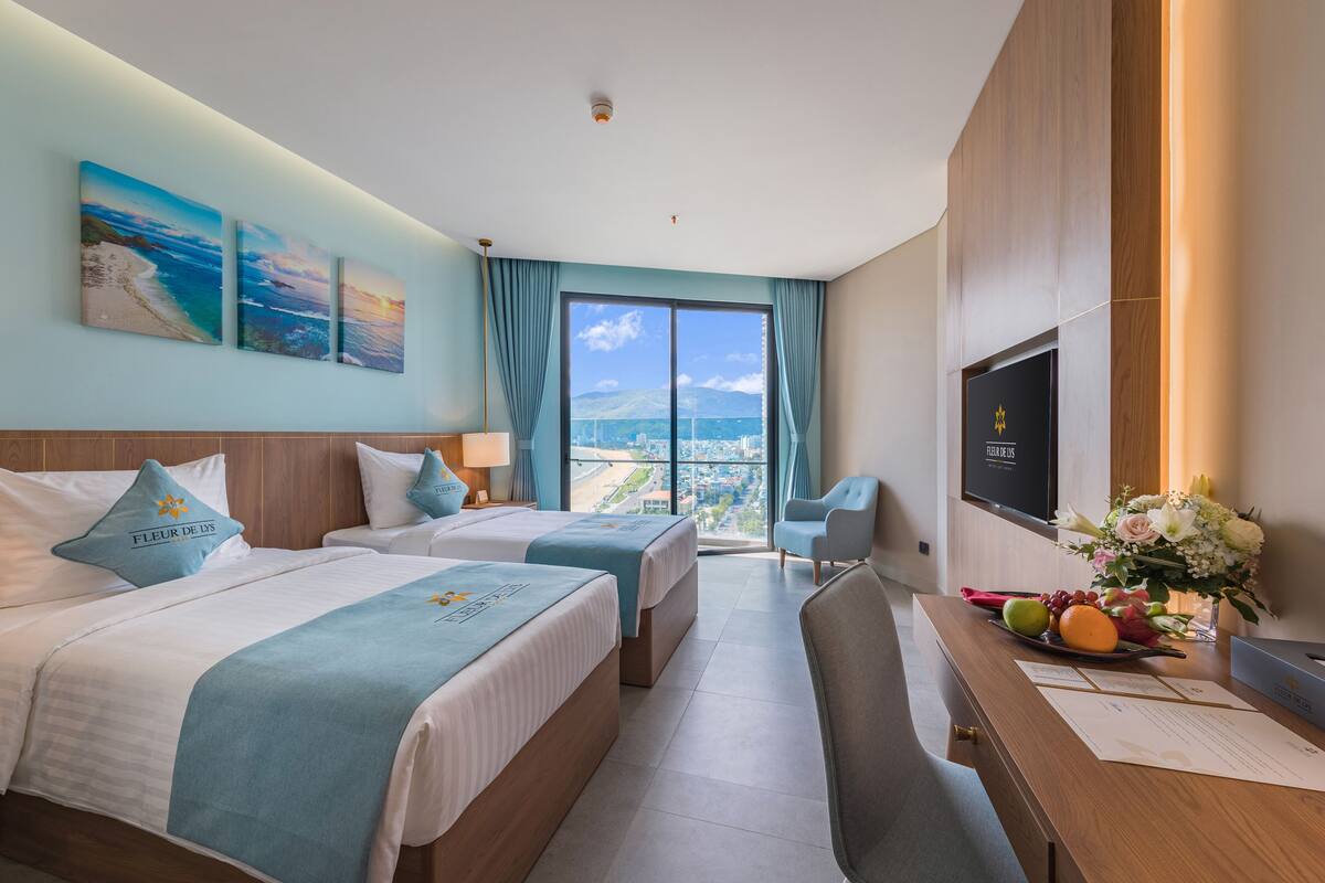 Habitación de hotel con dos camas, un escritorio, televisor y un amplio ventanal con vista a la ciudad.