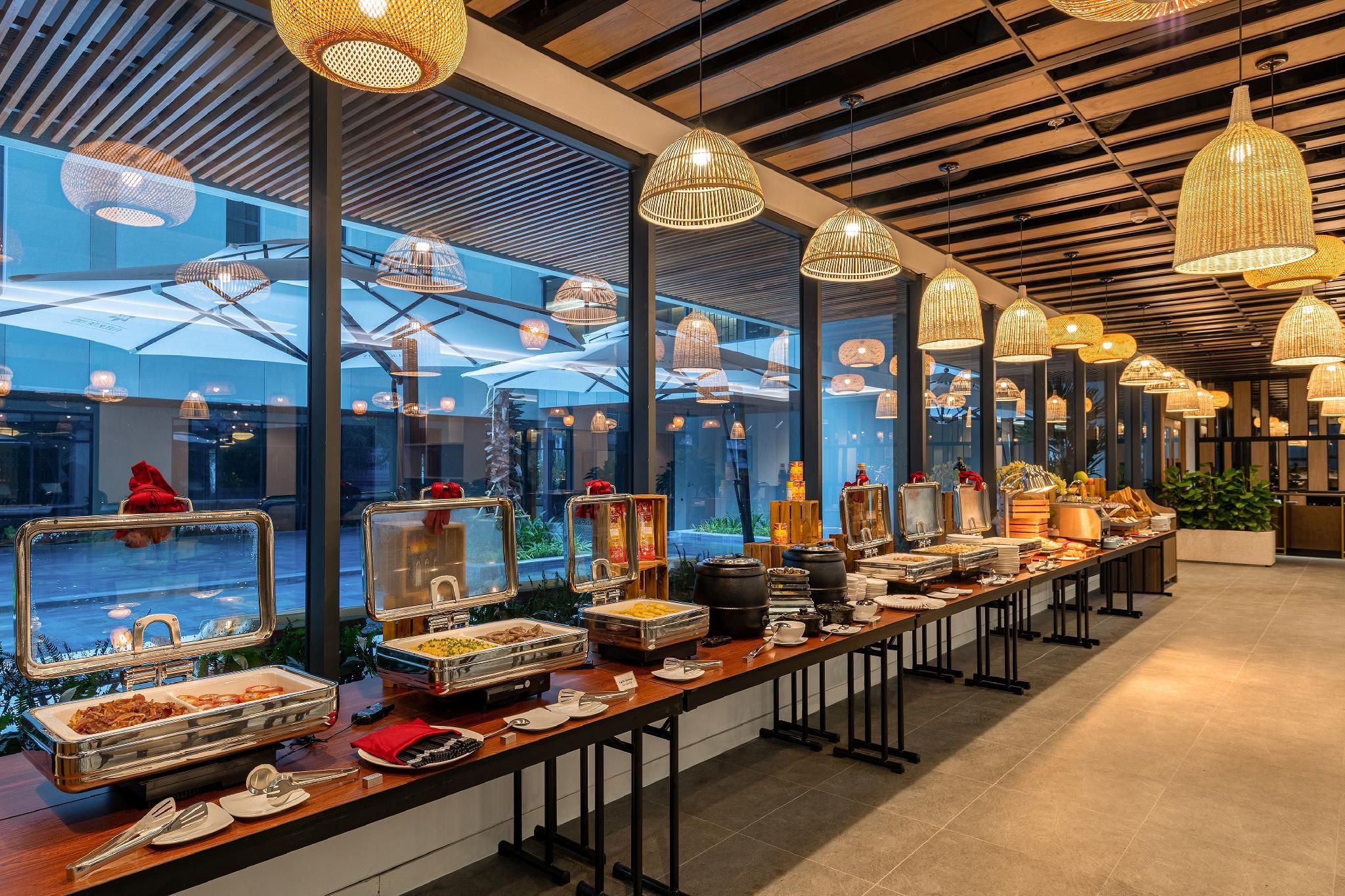 A buffet setup with various food items and utensils, large windows, and hanging pendant lights.