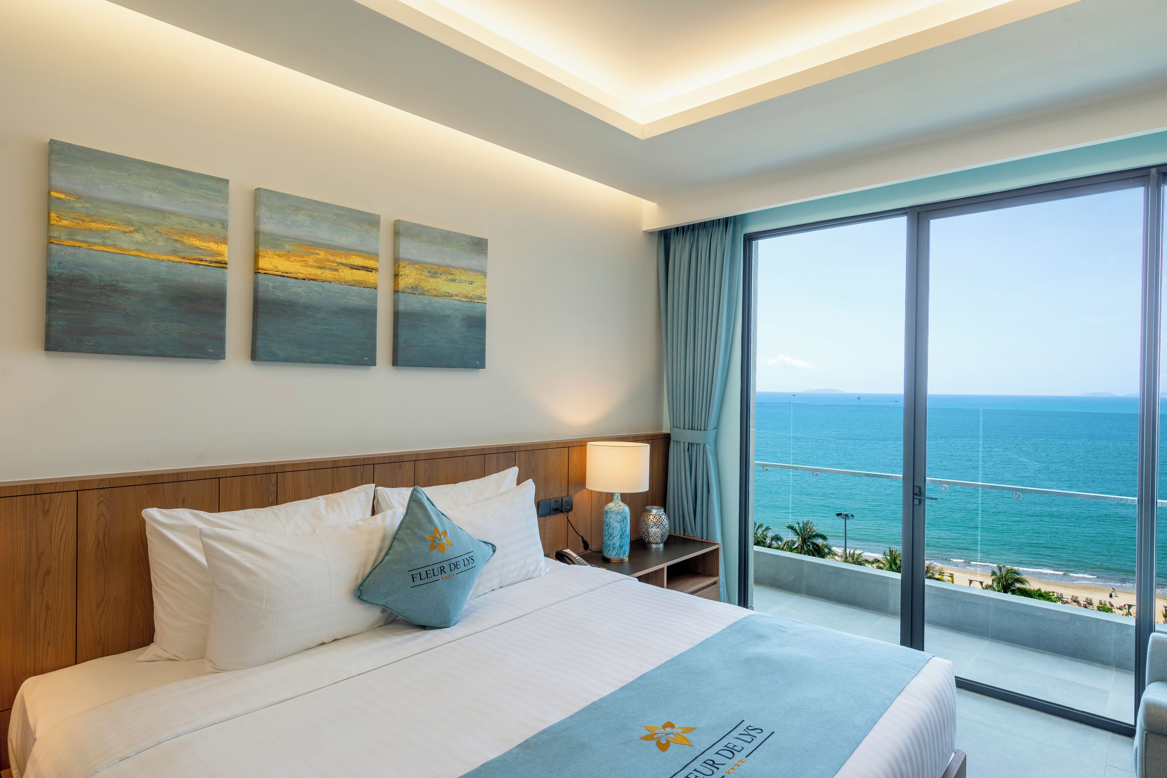 Quarto de hotel com uma cama grande, mesa de cabeceira, abajur e vista para o mar.