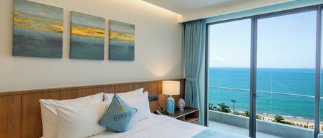 Quarto de hotel com uma cama grande, mesa de cabeceira, abajur e vista para o mar.
