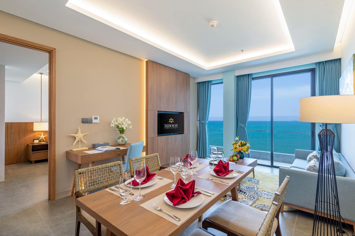 Habitación de hotel con comedor listo para cuatro personas, un sofá, un televisor y vista al mar.