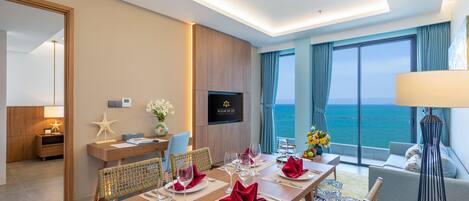 A hotel room with a dining area set for four, a sofa, a TV, and a view of the sea.