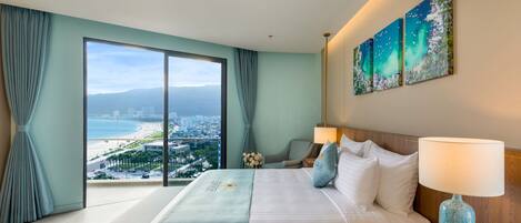 Quarto de hotel com uma cama grande, uma mesa de cabeceira, uma luminária e vista para uma praia e para o horizonte urbano através de uma janela ampla.