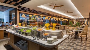 Un espace buffet avec différents stands de nourriture, un coin repas avec des tables et des chaises, et un plafond équipé de ventilateurs et de luminaires suspendus.