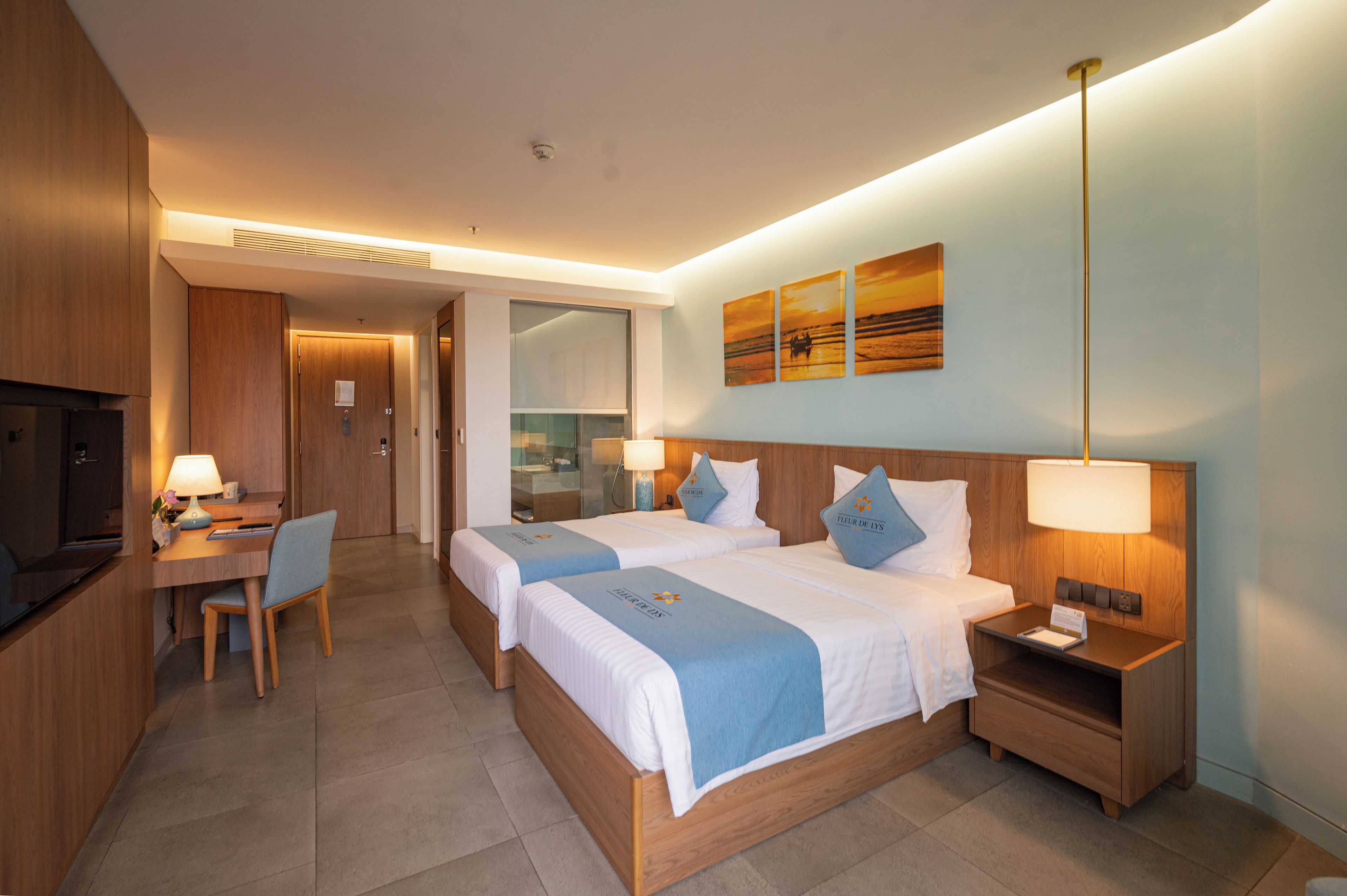 A hotel room with two beds, a desk, a chair, a TV, and artwork on the wall.