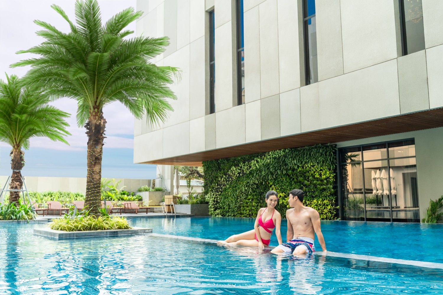 A swimming pool with two people sitting on the edge, surrounded by palm trees and a modern building with large windows.