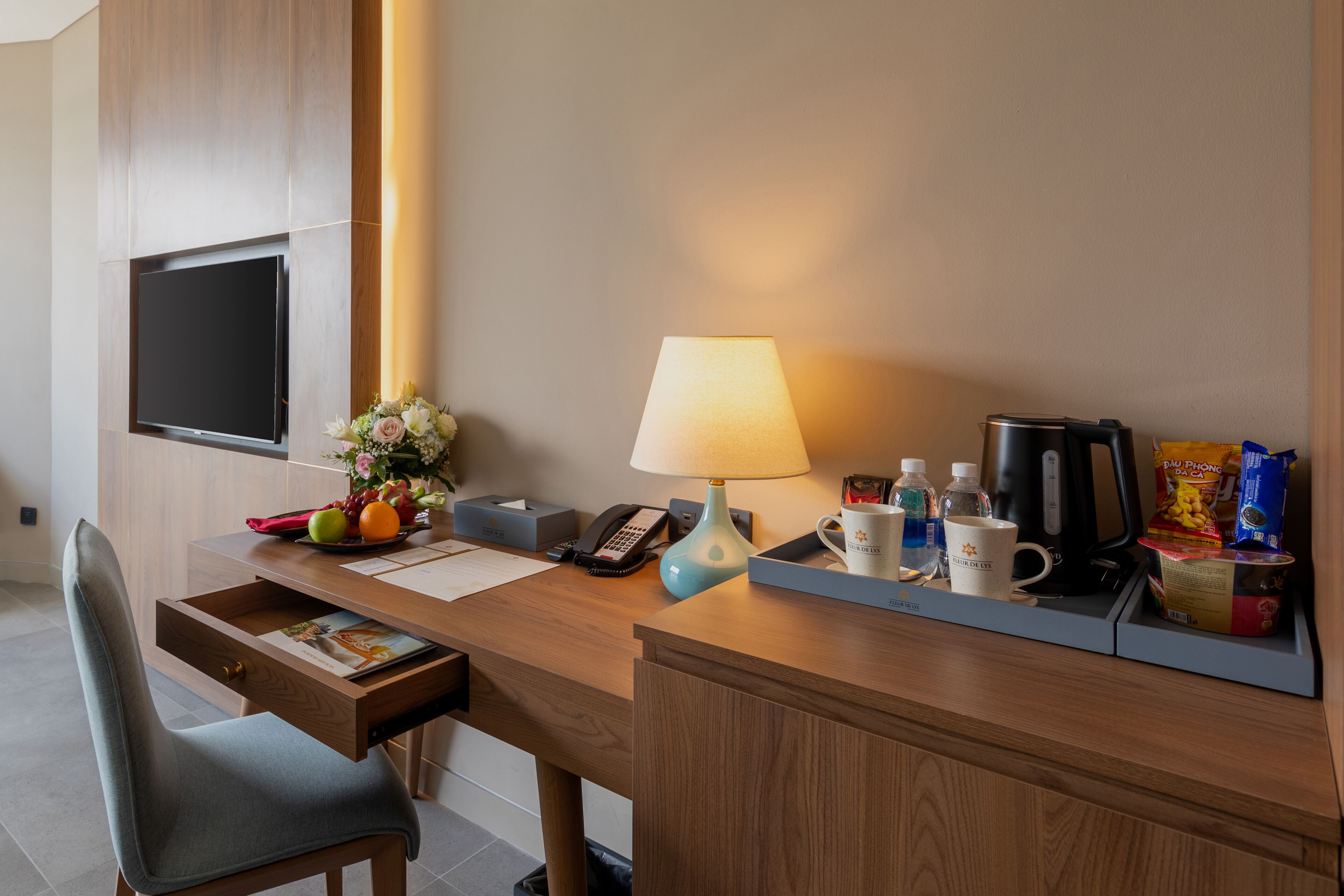 A hotel room with a wooden desk, a flat-screen TV mounted on the wall, a lamp, a telephone, a coffee maker, and various personal items.