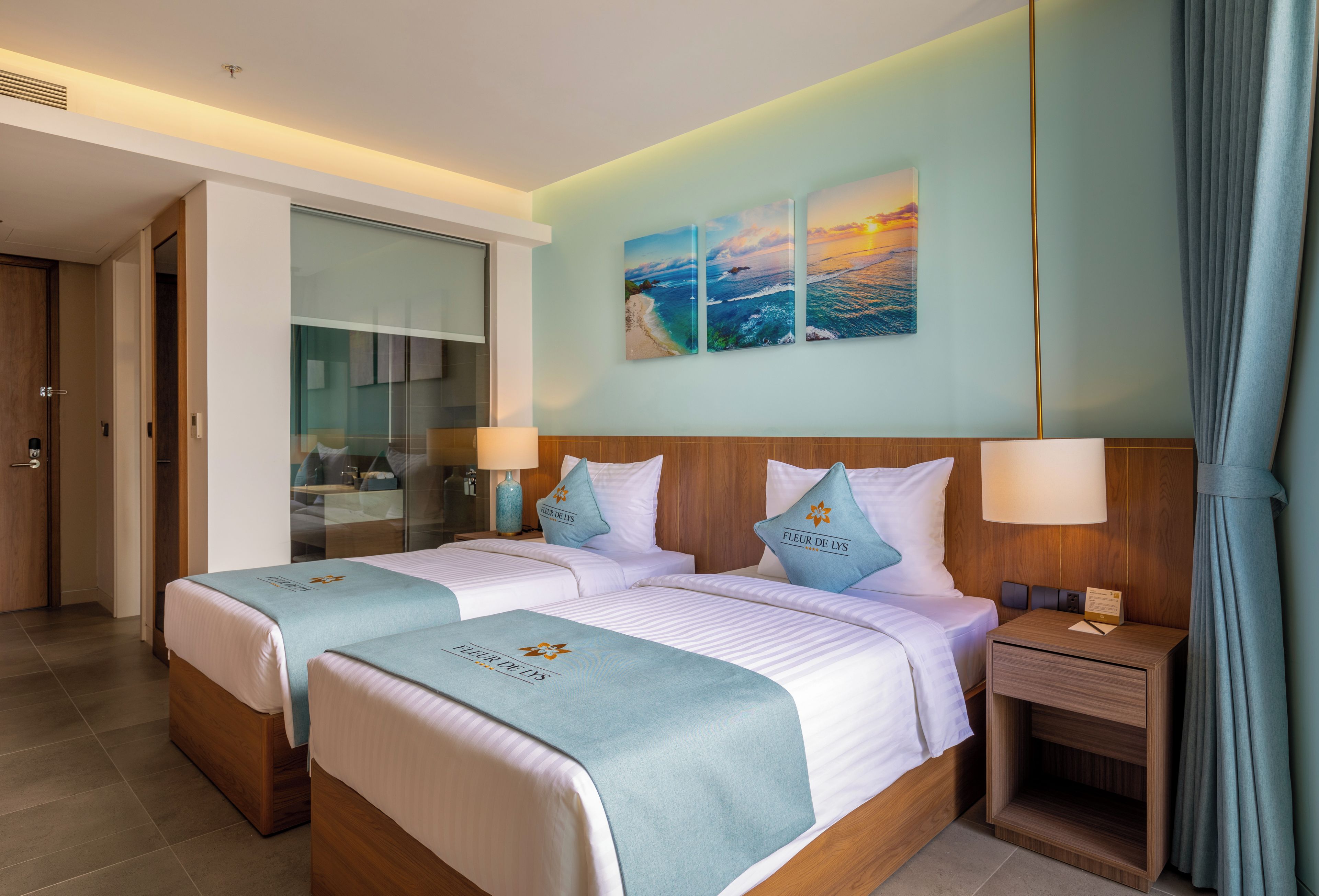 A hotel room with two beds, a large window, a painting of a beach scene, and a nightstand with a lamp.